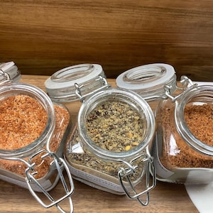 May include: Four glass jars with metal lids filled with different types of spices. The jars are arranged in a row on a wooden surface.
