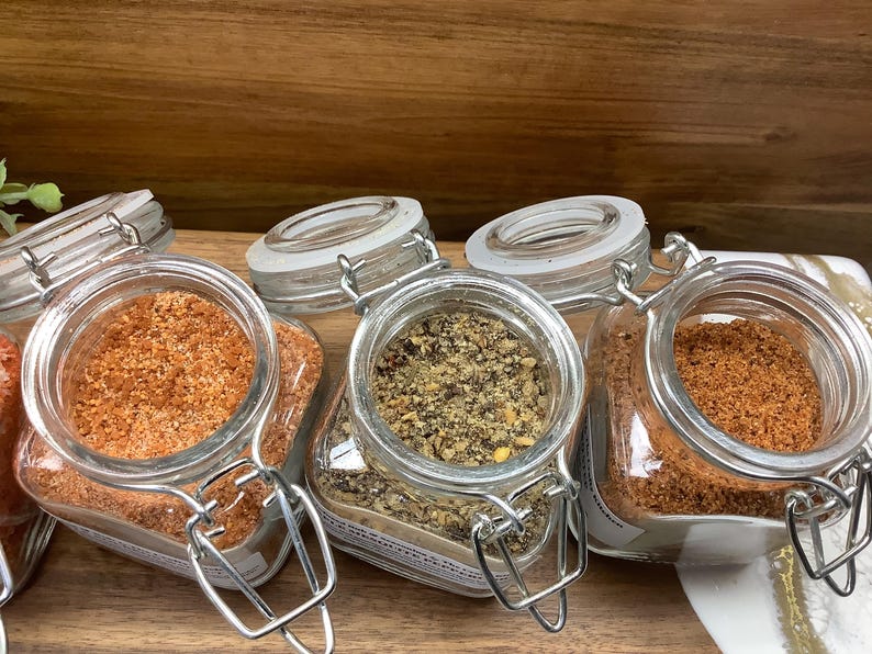 May include: Four glass jars with metal lids filled with different types of spices. The jars are arranged in a row on a wooden surface.