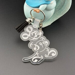 May include: A clear acrylic keychain featuring a white silhouette of Mickey Mouse. The keychain has a black tassel attached to a silver ring.