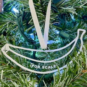 May include: A clear, banana-shaped Christmas ornament with the text "FOR SCALE" printed on it. The ornament hangs from a white ribbon against a backdrop of a green Christmas tree with blue lights.