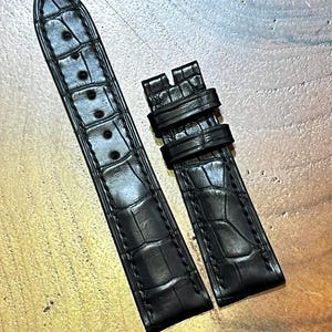 May include: Black watch strap with a crocodile skin effect. The strap has a textured surface and multiple adjustment holes. The buckle and strap are separate.