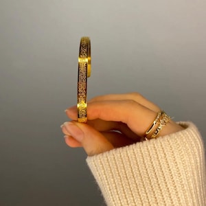 May include: A gold bangle bracelet with an intricate engraved design. The bracelet is open and has a simple, minimalist style.