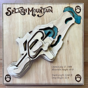 Puede incluir: Una pieza de arte de pared de madera que representa el paseo Splash Mountain en Disney World. La obra de arte está hecha de madera en capas y presenta un elemento de agua azul. El texto en la obra de arte dice "SPLASH MOUNTAIN", "Abierto el 17 de julio de 1989", "Altura de la montaña: 89 pies", "Longitud de la pista: 2.640 pies" y "Altura de la caída: 52 pies".