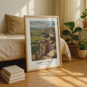 Könnte beinhalten: Ein gerahmter Kunstdruck mit einer Landschaftsillustration des Peak District Nationalparks. Das Bild zeigt felsige Klippen mit violettem Heidekraut im Vordergrund, die ein grünes Tal unter einem bewölkten Himmel überblicken. Der Text lautet PEAK DISTRICT NATIONAL PARK.
