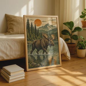 May include: A framed rectangular wall art print featuring a brown moose wading through a calm lake. The scene includes a dense forest of pine trees, a distant mountain range, and a large orange sun in the sky. The artwork is encased in a light wood frame.