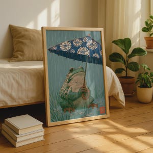 以下が含まれることがあります： 白い花柄の青い傘をさして雨の中に座る緑色のカエルの絵が描かれた額入りアートプリント。イラストには、草むらの中にいるカエルと、目に見える雨粒、そして隅にある小さな赤い四角いスタンプが描かれています。