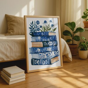 May include: A framed art print featuring a stack of five books with blue and white floral patterns on their covers. The books are arranged vertically, and a cluster of blue and white flowers and foliage emerges from the top of the stack.