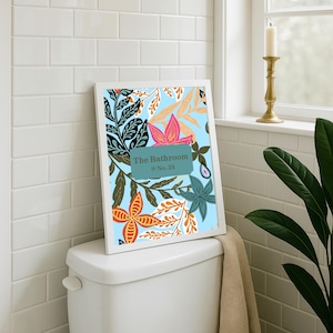 Può includere: Una stampa artistica incorniciata con un motivo floreale arancione, rosa e blu su sfondo azzurro. Il testo "The Bathroom @ No. 24" è visualizzato su uno striscione turchese. La stampa è appoggiata su un water bianco.
