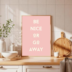 May include: A framed pink art print with the words "BE NICE OR GO AWAY" in white block letters. The print is displayed on a wooden countertop with a white ceramic bowl, vases, and a wooden cutting board.