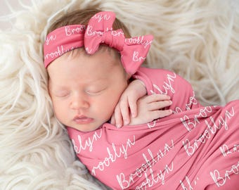 May include: A pink swaddle blanket with the name "Brooklyn" written in white script all over it. A baby girl is sleeping in the swaddle, wearing a pink headband with the same name written on it.