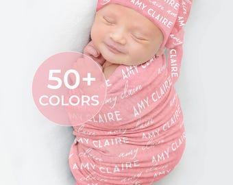 May include: A baby wrapped in a pink swaddle and matching hat, both printed with the name "Amy Claire" in white script. The image includes a pink circle with the text "50+ COLORS".