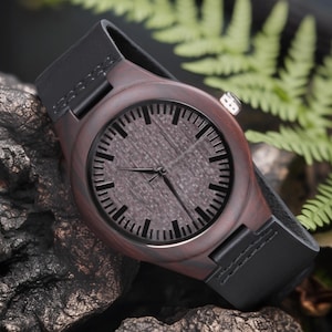 May include: A wooden wristwatch with a dark brown face and a black leather band. The watch face has black hour markers and hands. The watch is set against a dark, textured background with green foliage.