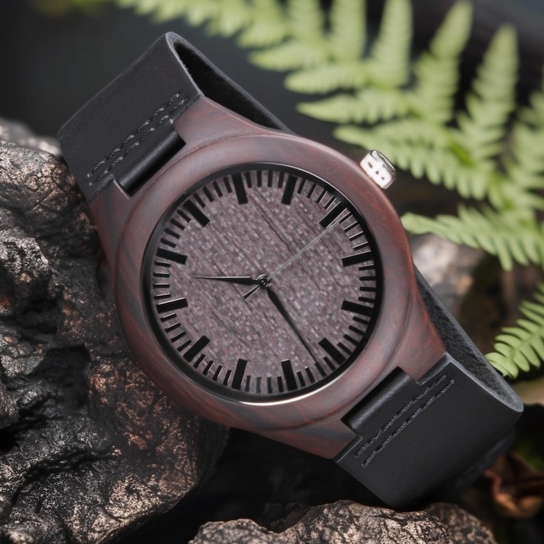 May include: A wooden wristwatch with a dark brown face and a black leather band. The watch face has black hour markers and hands. The watch is set against a dark, textured background with green foliage.