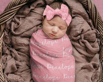 May include: A pink swaddle blanket with the name "Penelope" written in white script all over it. A baby is wrapped in the blanket and wearing a pink bow headband with the name "Penelope" written on it.