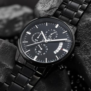 May include: A black wristwatch with a black metal band. The watch face has white markers and hands, with three sub-dials and a date display. The watch is set against a dark, textured background.
