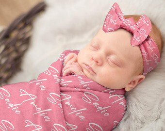 May include: A pink baby swaddle blanket with the name "Sadie" printed in white script. The blanket is wrapped around a sleeping baby wearing a pink headband with the name "Sadie" printed in white script.