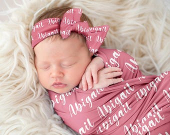 May include: A pink swaddle blanket with the name Abigail printed in white script. A matching pink headband with the name Abigail printed in white script is on the baby's head.