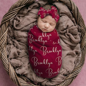 Pode incluir: Um rec&eacute;m-nascido envolto em um cobertor e faixa de cabelo bord&ocirc; combinando, ambos adornados com o nome "Brooklyn" em uma escrita delicada. O beb&ecirc; descansa pacificamente em uma cesta de vime forrada com tecido macio de tons neutros.