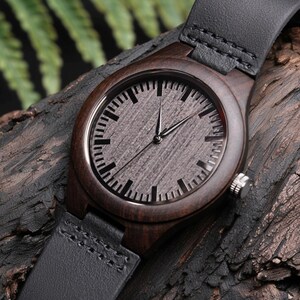 May include: A wooden wristwatch with a dark brown wood case and a gray face. The watch has black hands and a black leather band. The watch is displayed on a piece of dark wood.