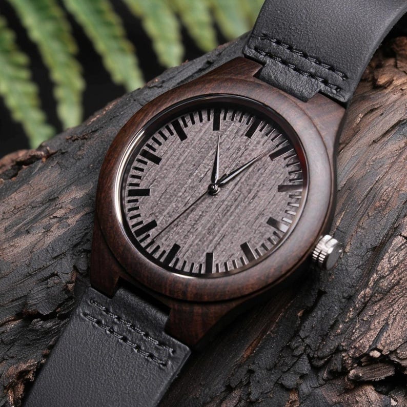 May include: A wooden wristwatch with a dark brown wood case and a gray face. The watch has black hands and a black leather band. The watch is displayed on a piece of dark wood.