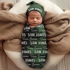 Pode incluir: Um rec&eacute;m-nascido envolto em um cobertor e gorro verde escuro, ambos com o nome "SAM JAMES" em escrita branca. O beb&ecirc; est&aacute; aninhado em um travesseiro branco, coberto por um cobertor marrom.