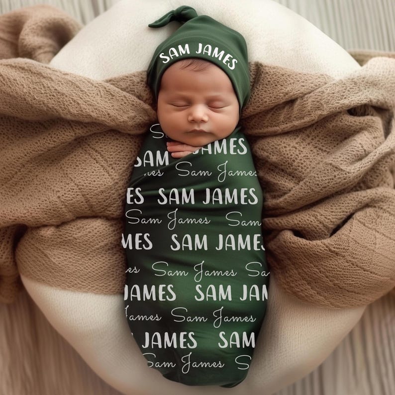 Pode incluir: Um rec&eacute;m-nascido envolto em um cobertor e gorro verde escuro, ambos com o nome "SAM JAMES" em escrita branca. O beb&ecirc; est&aacute; aninhado em um travesseiro branco, coberto por um cobertor marrom.