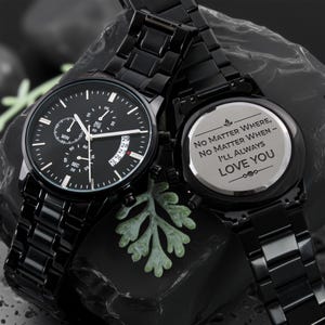 May include: Two black watches with matching bands. One watch face displays the time with multiple sub-dials and a date window. The other watch has the inscription "No Matter Where, No Matter When - I'll Always Love You."