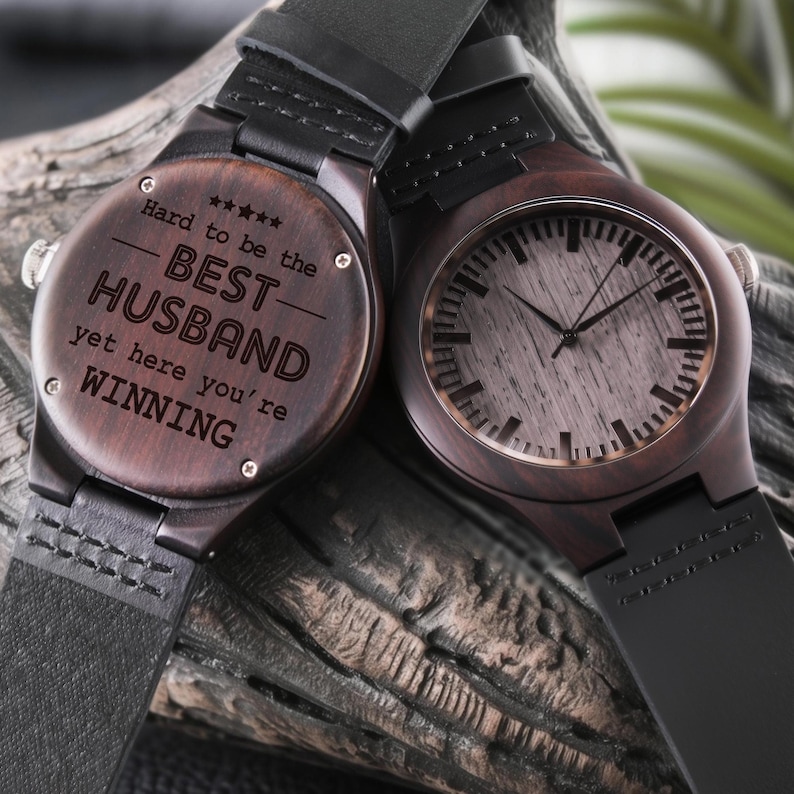 May include: Two wooden watches with black leather straps. One watch face has the text "Hard to be the BEST HUSBAND yet here you're WINNING". The other watch face has a light wood grain design. Both watches are on a wooden surface.