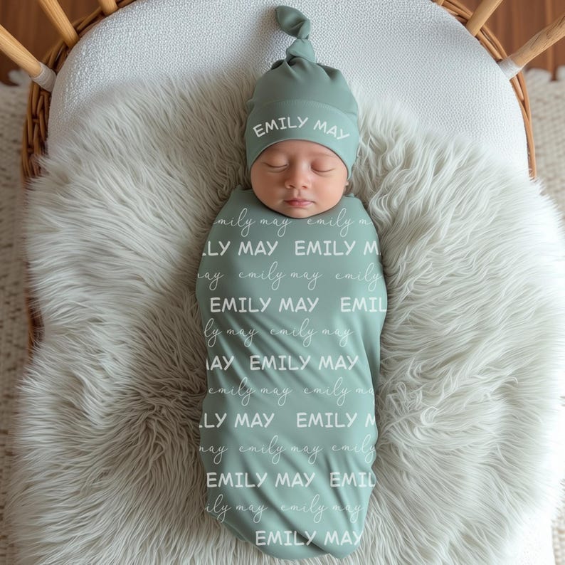 Pode incluir: Um beb&eacute; envolto numa manta e chap&eacute;u atado verde s&aacute;lvia a condizer. O nome "EMILY MAY" est&aacute; escrito em branco na manta e no chap&eacute;u. O beb&eacute; est&aacute; deitado numa superf&iacute;cie branca e fofa.
