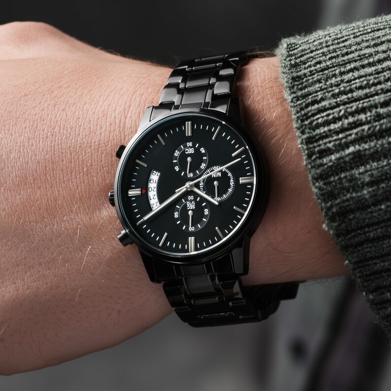 May include: A black wristwatch with a black metal band is displayed. The watch face has white markings and three smaller dials. The watch is on a wrist, with a gray sweater sleeve in the background.