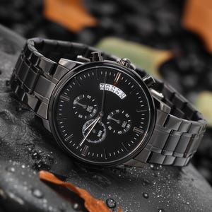 May include: A black wristwatch with a black metal band. The watch face has multiple sub-dials and white markings. The watch is set against a dark, textured background, with a few leaves visible.