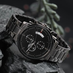 May include: A black wristwatch with a matching metal band. The watch face has multiple sub-dials and a date display. The watch is set against a dark, textured background, highlighting its sleek design.