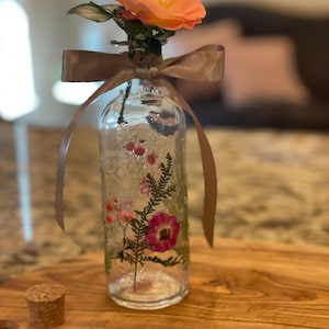 May include: A clear glass bottle with a brown ribbon tied around the neck. The bottle is filled with pressed flowers, including a pink rose and green leaves. The bottle is sitting on a wooden surface.