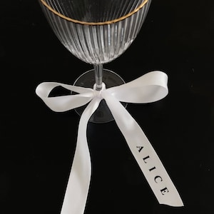 May include: A clear glass wine glass with a gold rim and a white ribbon tied around the stem. The ribbon has the name "Alice" printed on it.
