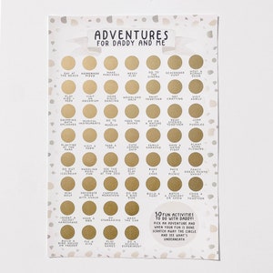 May include: A white card with gold circles and black text. The card has a title that says "Adventures for Daddy and Me". The card has a list of 50 fun activities to do with your dad.  The card says "Pick an adventure and when your fun is done scratch away the circle and see what's underneath."