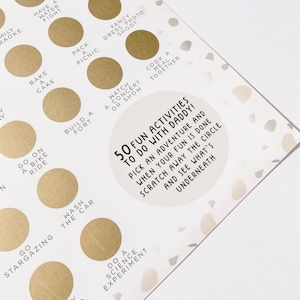 May include: A gold and white game board with 50 fun activities to do with daddy. The board has a circle with a scratch-off surface. The text on the board says "50 Fun Activities to Do With Daddy! Pick an adventure and when your fun is done scratch away the circle and see what's underneath."