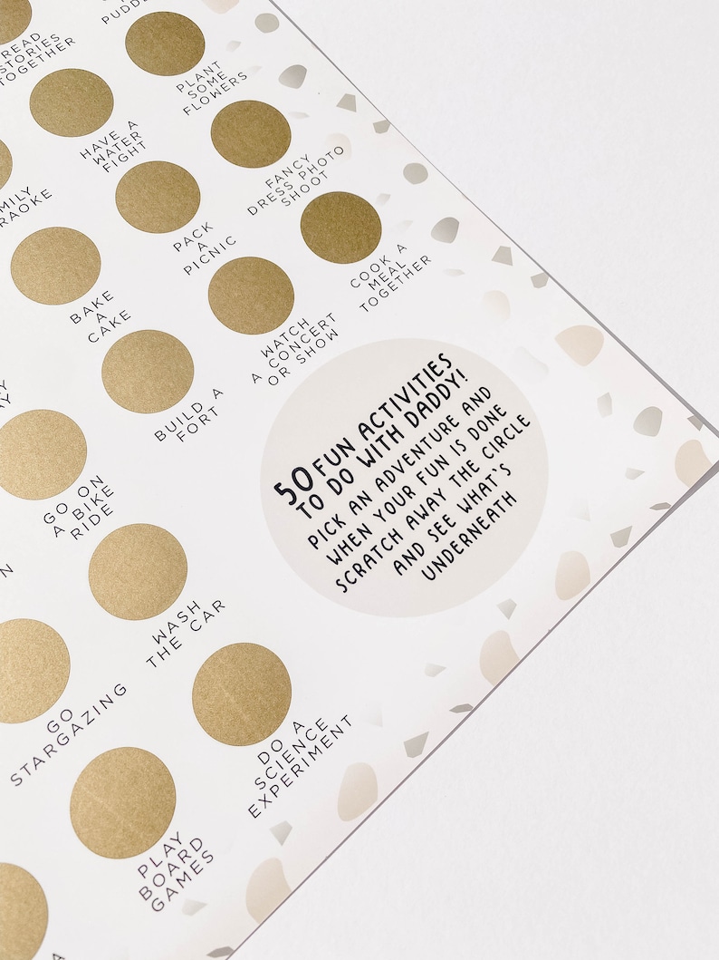May include: A gold and white game board with 50 fun activities to do with daddy. The board has a circle with a scratch-off surface. The text on the board says "50 Fun Activities to Do With Daddy! Pick an adventure and when your fun is done scratch away the circle and see what's underneath."