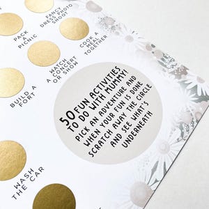 Op de afbeelding: Een witte kaart met een gouden cirkel in het midden. De cirkel heeft de tekst "50 Fun Activities to do with Mummy!"  Kies een avontuur en wanneer je klaar bent met je plezier, schraap de cirkel weg en zie wat eronder zit. De kaart is omringd door een wit madeliefjespatroon.