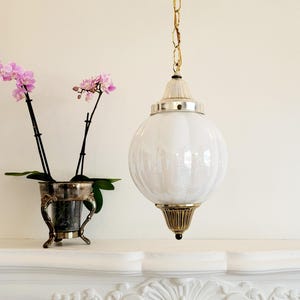 May include: A white globe pendant light fixture with a gold chain and a gold ceiling mount. The light fixture is hanging above a white fireplace mantel.