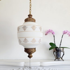 May include: Vintage white and gold pendant light fixture. The glass shade features a gold floral pattern and is suspended by a gold chain. The fixture has ornate gold metal accents and a pull chain. The light hangs above a white mantel.