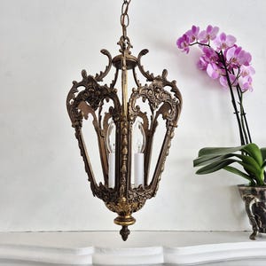 Puede incluir: Lámpara de araña de latón ornamentada con un diseño calado decorativo. La lámpara tiene dos bombillas en forma de vela y está suspendida por una cadena. Una orquídea en maceta con flores moradas está en el fondo.