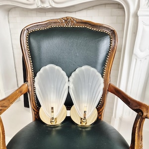May include: Two Art Deco-style wall sconces with a frosted glass shell design. The sconces have a gold-colored base and a vertical support. They are displayed on a dark green leather chair with a wooden frame.
