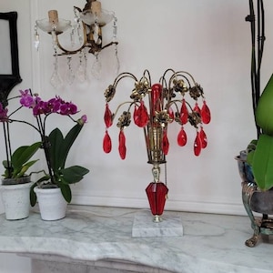 Puede incluir: Una lámpara de mesa dorada con una pantalla de cristal rojo y gotas de cristal rojo que cuelgan de la base. La lámpara está sobre una superficie de mármol blanco con dos plantas de orquídeas en maceta en el fondo.