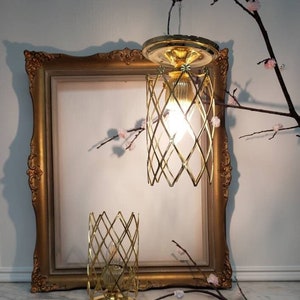 May include: Two gold caged light fixtures with a diamond pattern. The larger fixture is hanging from the ceiling and the smaller fixture is on a table. Both fixtures have a light bulb inside.