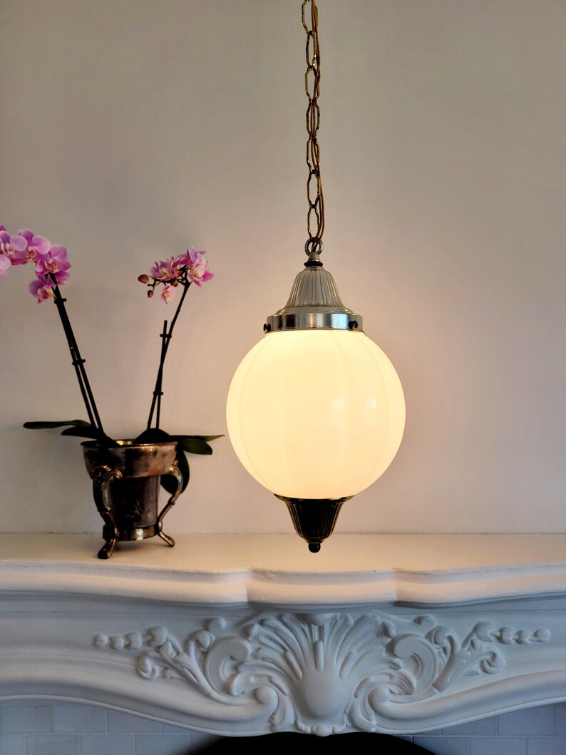 May include: A white frosted glass globe pendant light fixture with a silver chain and a silver and brass ceiling mount. The light is hanging above a white mantel with ornate carvings.