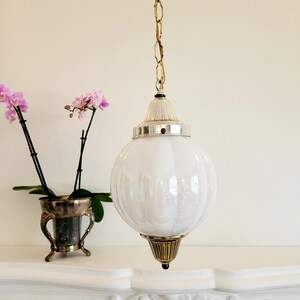 May include: A white glass globe pendant light fixture with a gold chain and a brass finial. The globe has a textured surface and is hanging from a white ceiling.