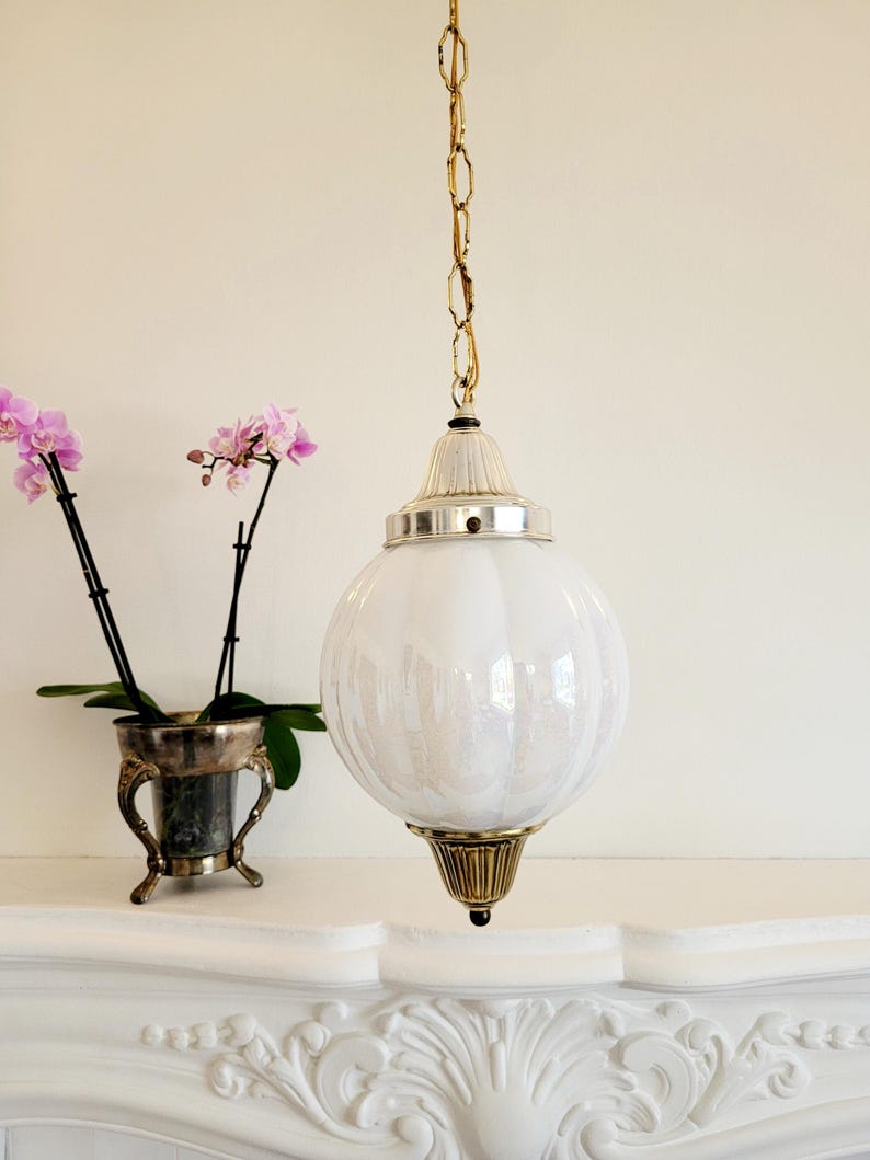 May include: A white glass globe pendant light fixture with a gold chain and a brass finial. The globe has a textured surface and is hanging from a white ceiling.