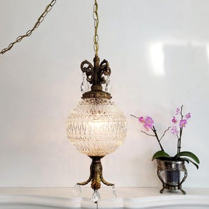 May include: A vintage brass and crystal chandelier with a faceted glass globe shade and crystal drops hanging from the bottom. The chandelier is hanging from a chain and is lit with a light bulb. The chandelier is in front of a white fireplace mantel.