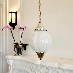 May include: A white glass globe pendant light fixture with a gold chain and a gold metal base. The light fixture is hanging from the ceiling in a room with white walls and a white fireplace.