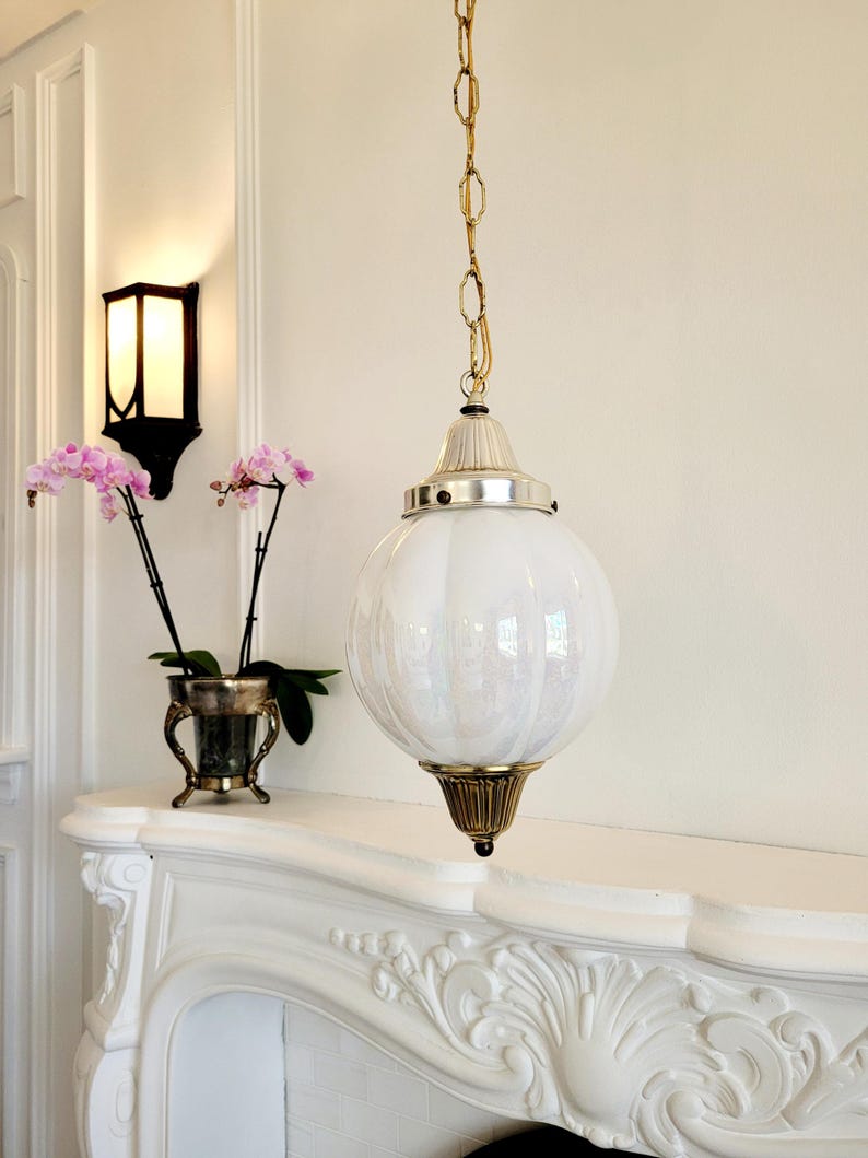 May include: A white glass globe pendant light fixture with a gold chain and a gold metal base. The light fixture is hanging from the ceiling in a room with white walls and a white fireplace.